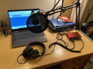 Photo of a desk with a laptop computer, a microphone on a boom with a breath shield on it, headphones, and related connection and storage gear commonly used for podcasting.