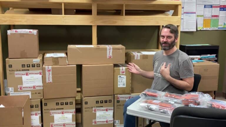 A photo of Drew gesturing towards a stack of cardboard boxes located behind him.