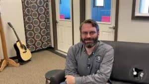 Drew smiling and talking while seated on a brown leather or vinyl couch