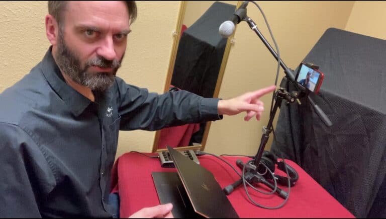 Photo of Drew pointing to a microphone on a boom attached to a table set up for podcasting with a laptop computer and other equipment
