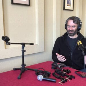 Photo of Drew wearing a black hoodie and shirt sitting behind a table with a red cloth cover with various podcast items displayed on the table in front of him