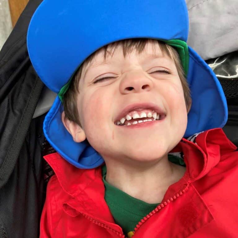 A close-up photo of a boy with short light-brown hair grinning broadly and wearing a bright blue bomber's hat and red coat.
