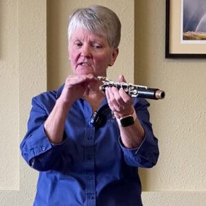 A photo of a woman with short gray hair and wearing a blue button-down shirt gesturing at a part of a clarinet while talking.