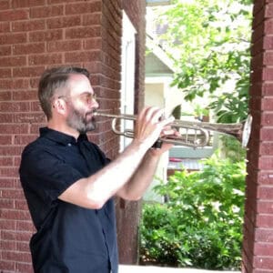 Profile photo of Drew playing the trumpet on the front porch of a house with a red brick exterior and lush green bushes and trees in the background.