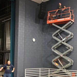 A photo of a man on a mechanical lift working on some hanging stage speakers
