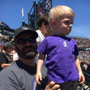 Photo of a man with a dark beard wearing a baseball cap with a small blonde boy standing on his lap in the stands at a Colorado Rockies baseball game