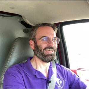 Photo of Drew smiling while driving a car with a gray and white interior and wearing a bright purple polo shirt.